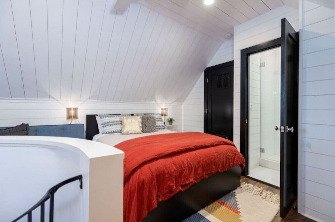 Un dormitorio con cama curvada, colcha roja, cabecera blanca, mesita de noche con lámpara y puerta corrediza de vidrio que da a una ducha.