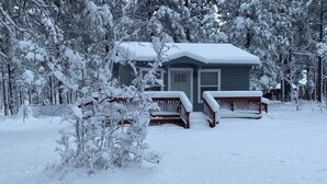 Una cabana amb un teulat cobert de neu, bancs de fusta i un paisatge nevós al voltant.