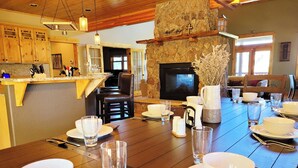A dining area with a stone fireplace, wooden furniture, and a kitchen in the background. - Mountain of Joy, 21-ZONE3029 (Estes Park)