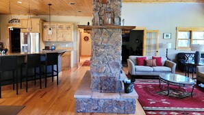 A spacious living area with a stone fireplace, wooden ceiling, and a kitchen with wooden cabinets and a stainless steel refrigerator. - Mountain of Joy, 21-ZONE3029 (Estes Park)