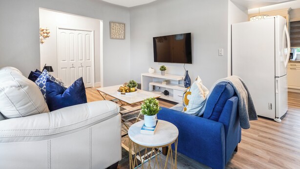 A modern living room with a white sofa, blue armchair, and a coffee table with a plant. A flat-screen TV is mounted on the wall, and a white refrigerator is visible in the background. - Urban Nest 7 mins from Edmonton downtown <br> (Edmonton)