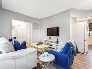 A modern living room with a white sofa, blue armchair, and a coffee table with a plant. A flat-screen TV is mounted on the wall, and a white refrigerator is visible in the background. - Urban Nest 7 mins from Edmonton downtown <br> (Edmonton)