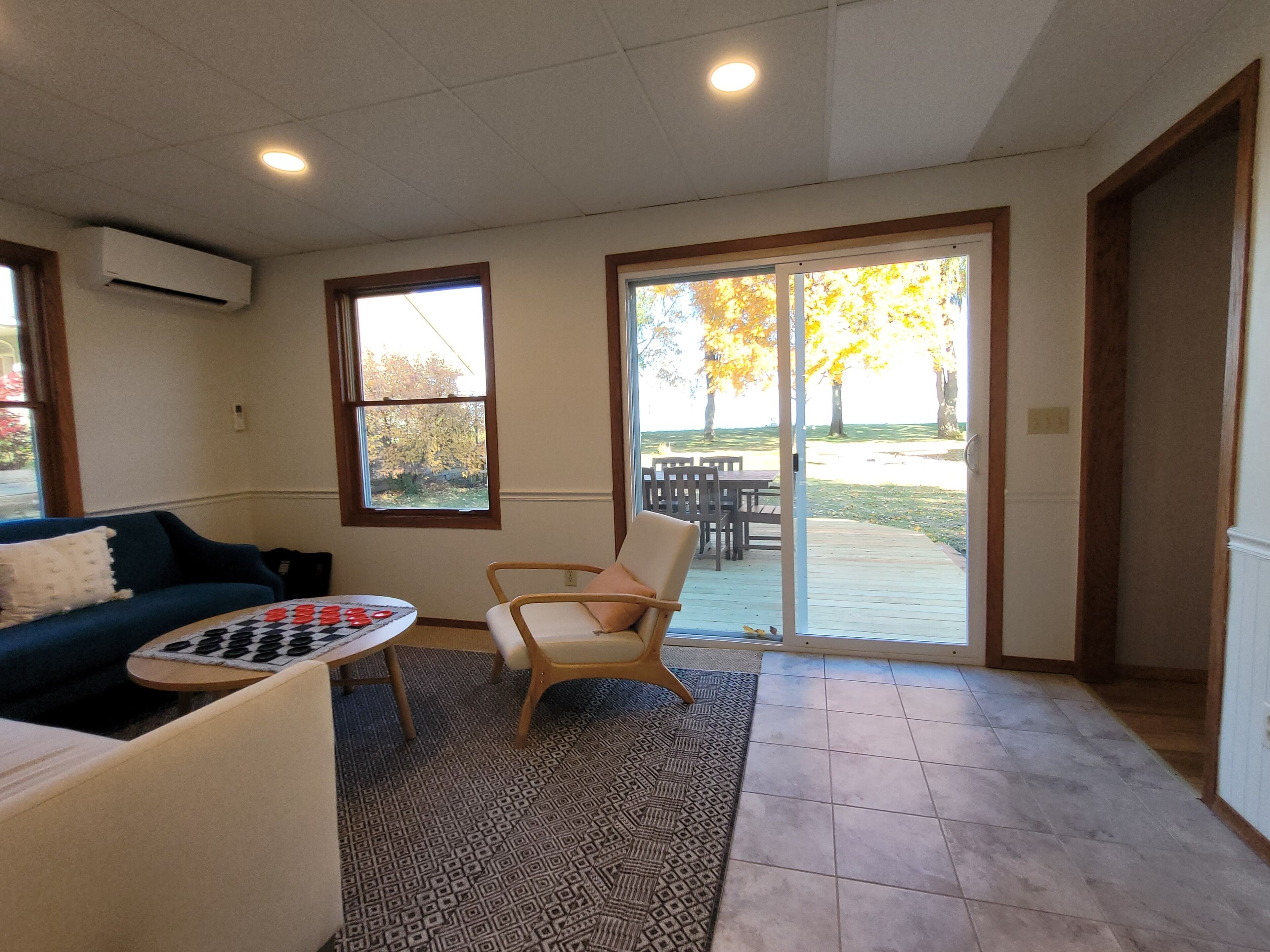 A spacious living room with a blue couch, a wooden chair, a patterned rug, and a table with a board game. There are large windows and a sliding glass door leading to an outdoor area with a patio and trees.