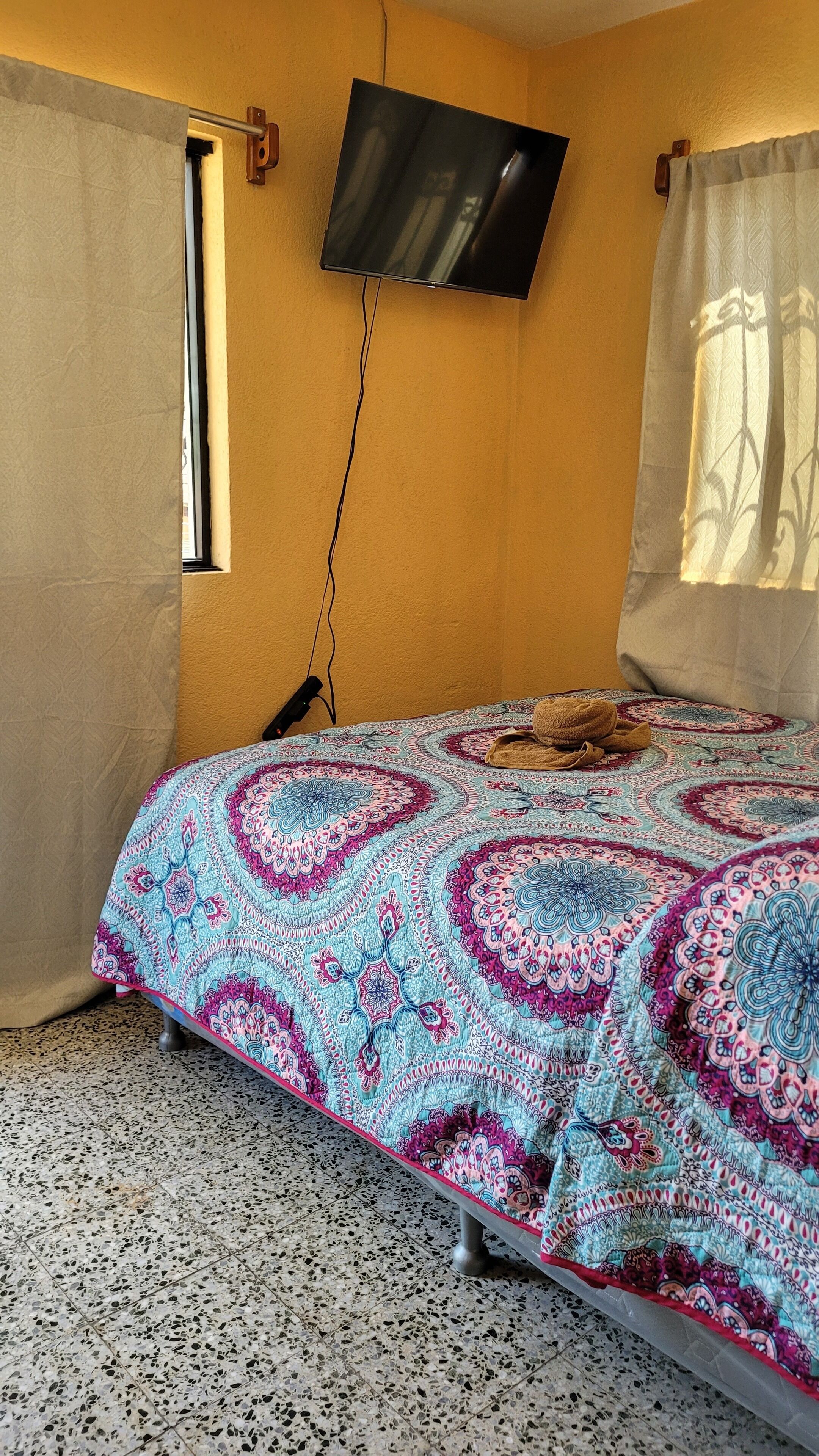 A bedroom with a patterned bedspread, a television mounted on the wall, and a window with curtains.