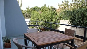 Un balcón con una mesa de madera y sillas de mimbre, con vistas a un jardín con plantas y árboles diversos.