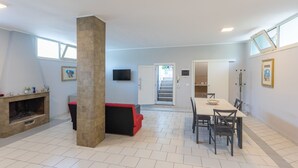 Amplia sala de estar con chimenea, un pilar de piedra, una mesa de comedor con sillas y una escalera que conduce a otra habitación.