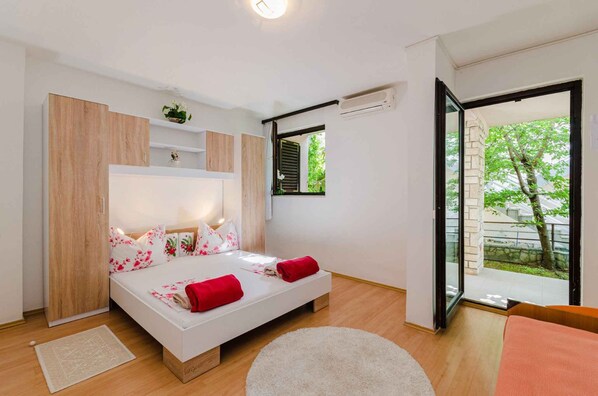 A bedroom with a bed, a wardrobe, a small shelf, a window, and a door leading to an outdoor area.