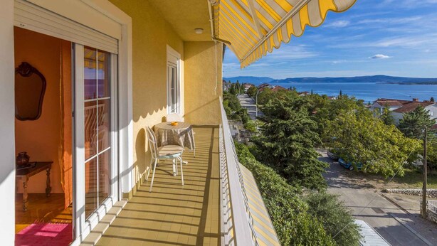 Un balcon agrémenté d’un auvent jaune à rayures, une table avec une chaise, et une vue sur une étendue d’eau et des maisons au loin.