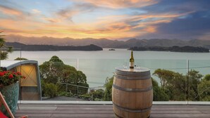 A scenic view from a balcony overlooking a lake with mountains in the background, a wooden barrel, and a glass enclosure with plants.