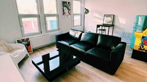 A living room with a black leather sofa, a white armchair, and a coffee table. There are three windows providing natural light, a floor lamp, and a framed picture on the wall. - London Flat Next to Central Line Tube Station (London)