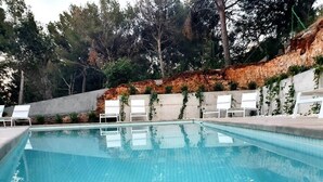 A swimming pool with clear blue water, surrounded by white lounge chairs and a concrete wall with greenery. - Apartamento con Piscina 1 C en Playa de Canyamel (Capdepera)