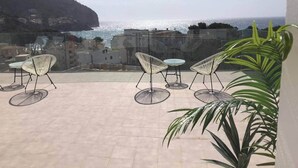 An outdoor terrace with chairs and tables, overlooking a coastal area with buildings and a hill. - Apartamento con Piscina 1 C en Playa de Canyamel (Capdepera)