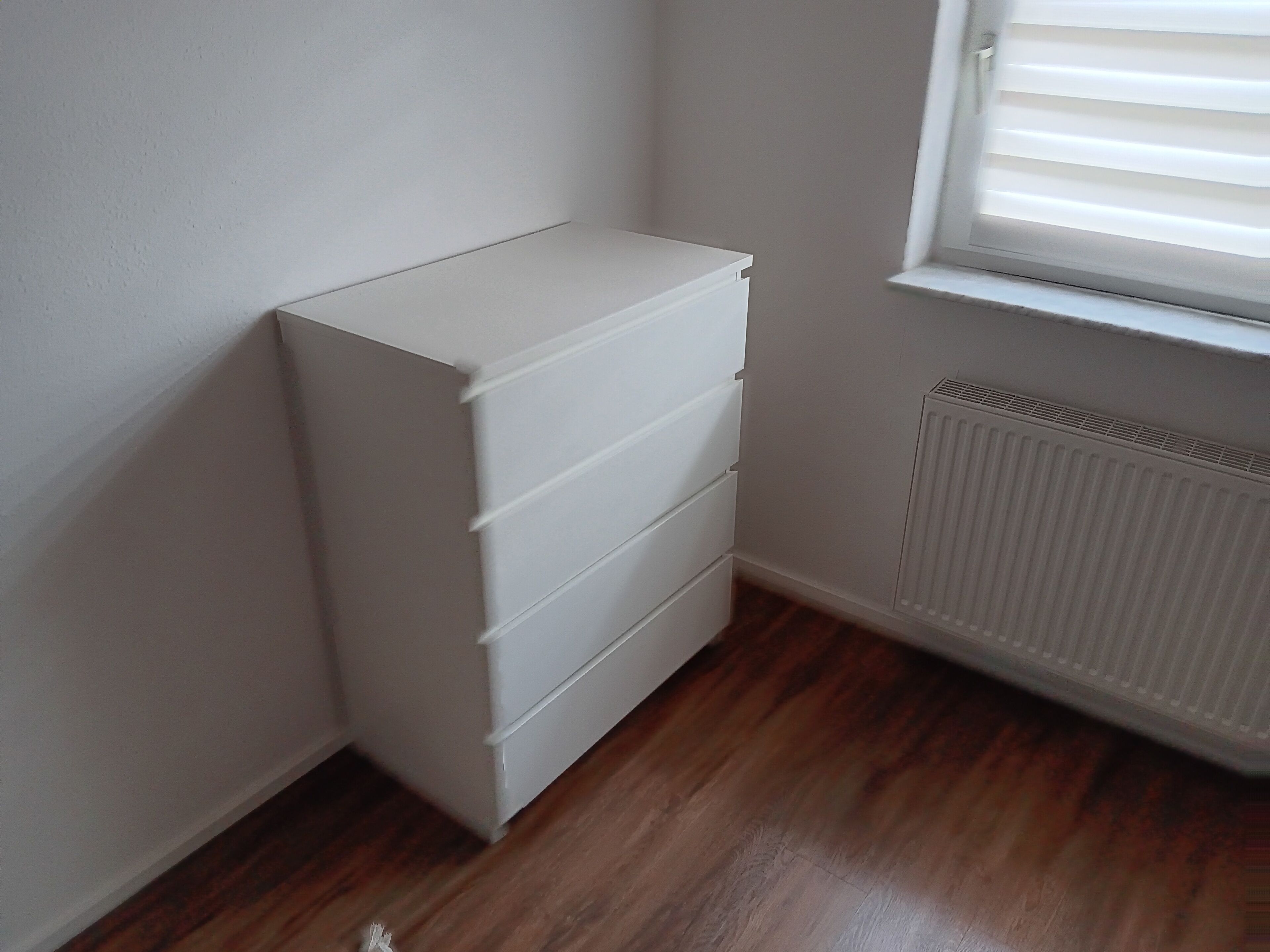 Ein Eckzimmer mit weißem Schminktisch, Holzboden und einem Fenster mit Jalousien.