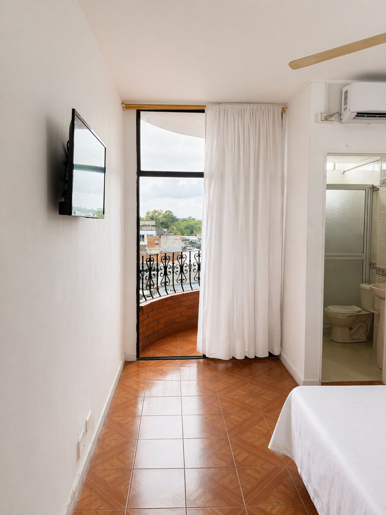 A room with a TV mounted on the wall, a bed, a bathroom with a toilet and sink, and a balcony with a view of buildings and greenery.