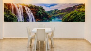 Uma área de refeições com mesa e cadeiras, um quadro grande de uma cascata e paisagem na parede, e iluminação embutida no teto.