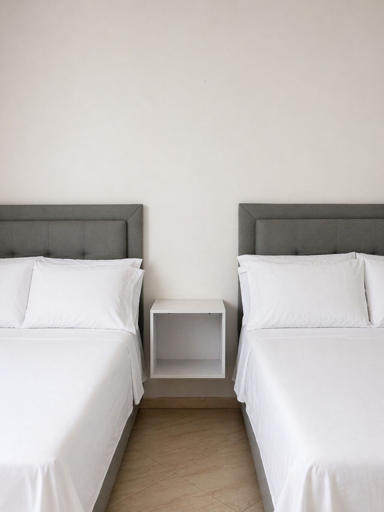 A twin room with two beds, a small white nightstand, and a plain wall.