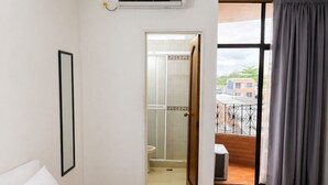Quarto de hotel com cama, unidade de ar condicionado, banheiro com chuveiro e varanda com vista para edifícios.
