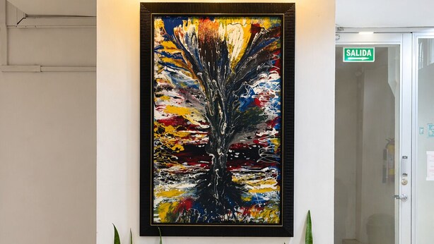 Um quadro abstrato emoldurado numa parede, três vasos de plantas por baixo e uma porta de vidro com o letreiro 'SALIDA'.