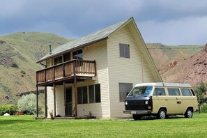 A single-story house with a balcony, a van parked in front, and a hilly landscape in the background. - Goldbug Hot Springs Getaway, Cozy Cabin Great for Hot Springs, Fishing & Hunting! (Salmon)