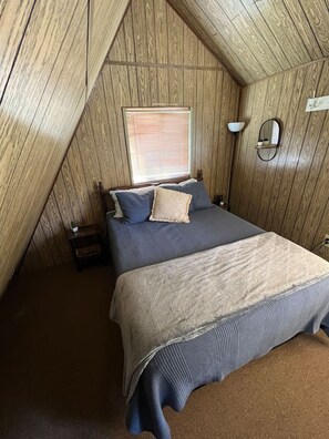 A bedroom with a wooden sloped ceiling, a bed with blue bedding, a window with blinds, and a bedside table. - Goldbug Hot Springs Getaway, Cozy Cabin Great for Hot Springs, Fishing & Hunting! (Salmon)