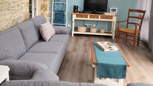Una sala de estar con un sofá gris, una silla de madera, un televisor sobre un mueble blanco y un tragaluz arriba.