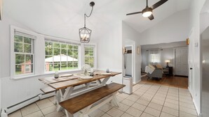 A spacious dining area with a wooden table and bench, large windows with a view of greenery, and a ceiling fan. - Peaceful Oasis minutes to T.F. Green International Airport, Main Street & more (East Greenwich)