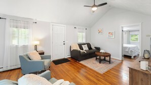 A spacious living room with a wooden floor, a grey sofa, a blue armchair, a small round table, and a ceiling fan. - Peaceful Oasis minutes to T.F. Green International Airport, Main Street & more (East Greenwich)