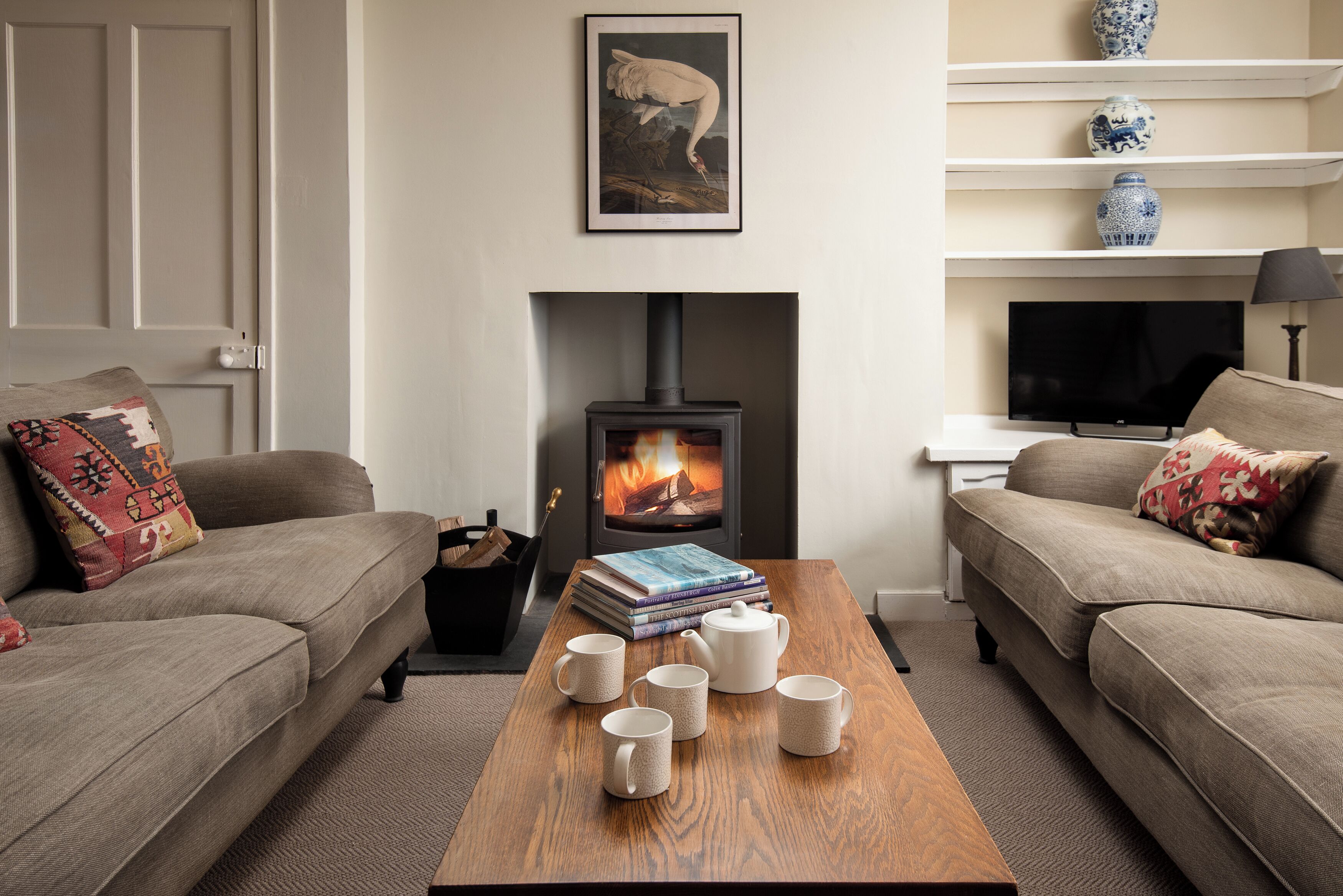 A cozy living room with a fireplace, sofas, a coffee table with books and mugs, and a TV.
