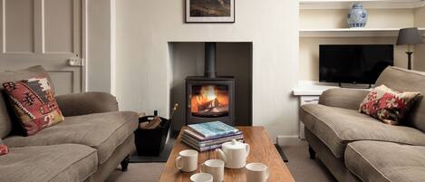 A cozy living room with a fireplace, sofas, a coffee table with books and mugs, and a TV.