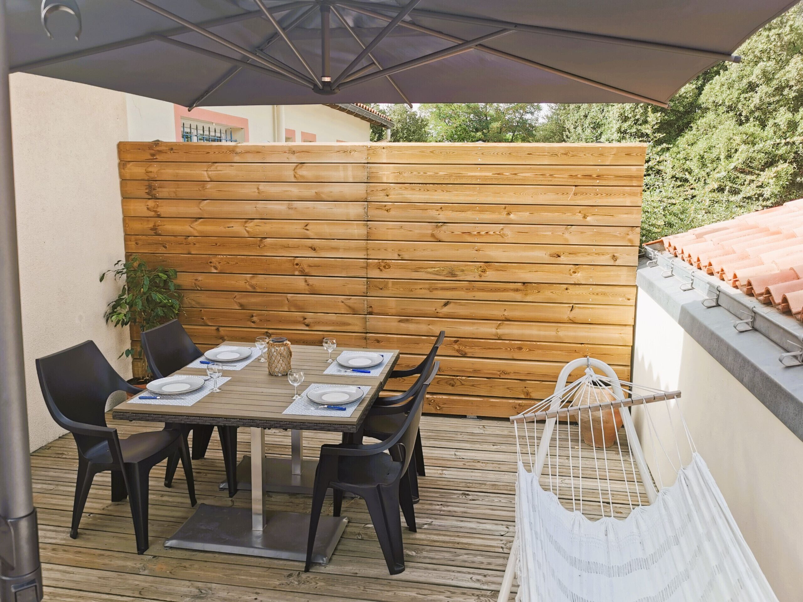 An outdoor patio with a wooden table set for four, surrounded by black chairs, under a canopy.