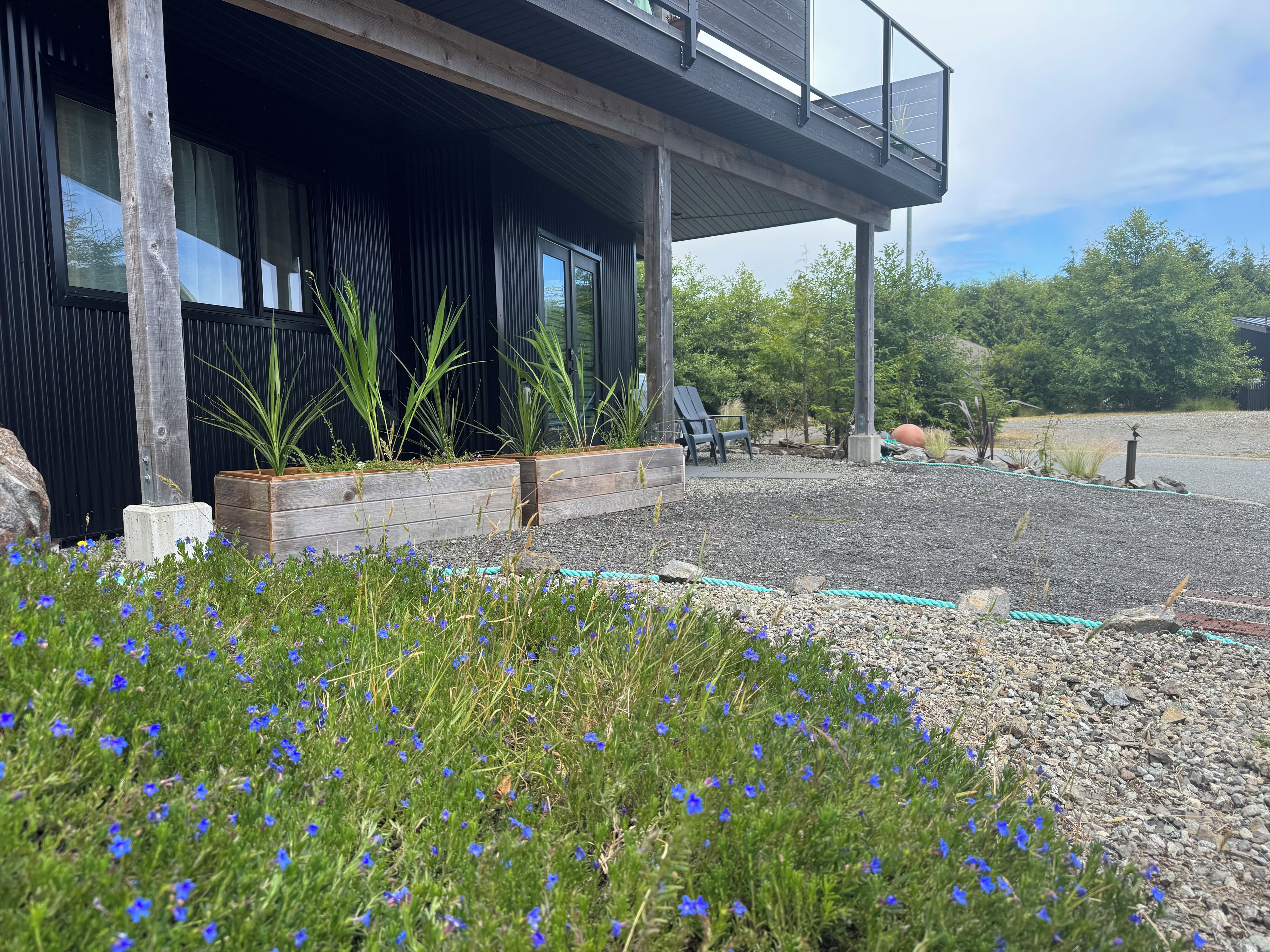 Ein modernes Gebäude mit einer überdachten Außenbereich-Sitzgelegenheit, einem Garten mit blauen Blumen und einem Kiesweg.