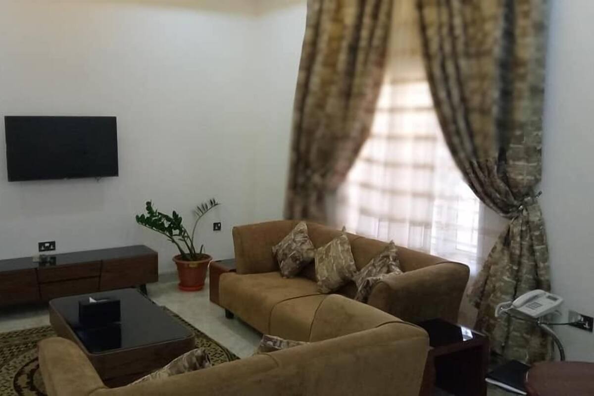 A living room with a sofa, a coffee table, and a TV mounted on the wall. There are potted plants and a telephone on a stand.