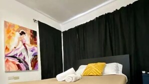 Moderni hotellihuone, jossa on suuri sänky, mustat verhot, taulu seinällä ja keltainen tyyny.