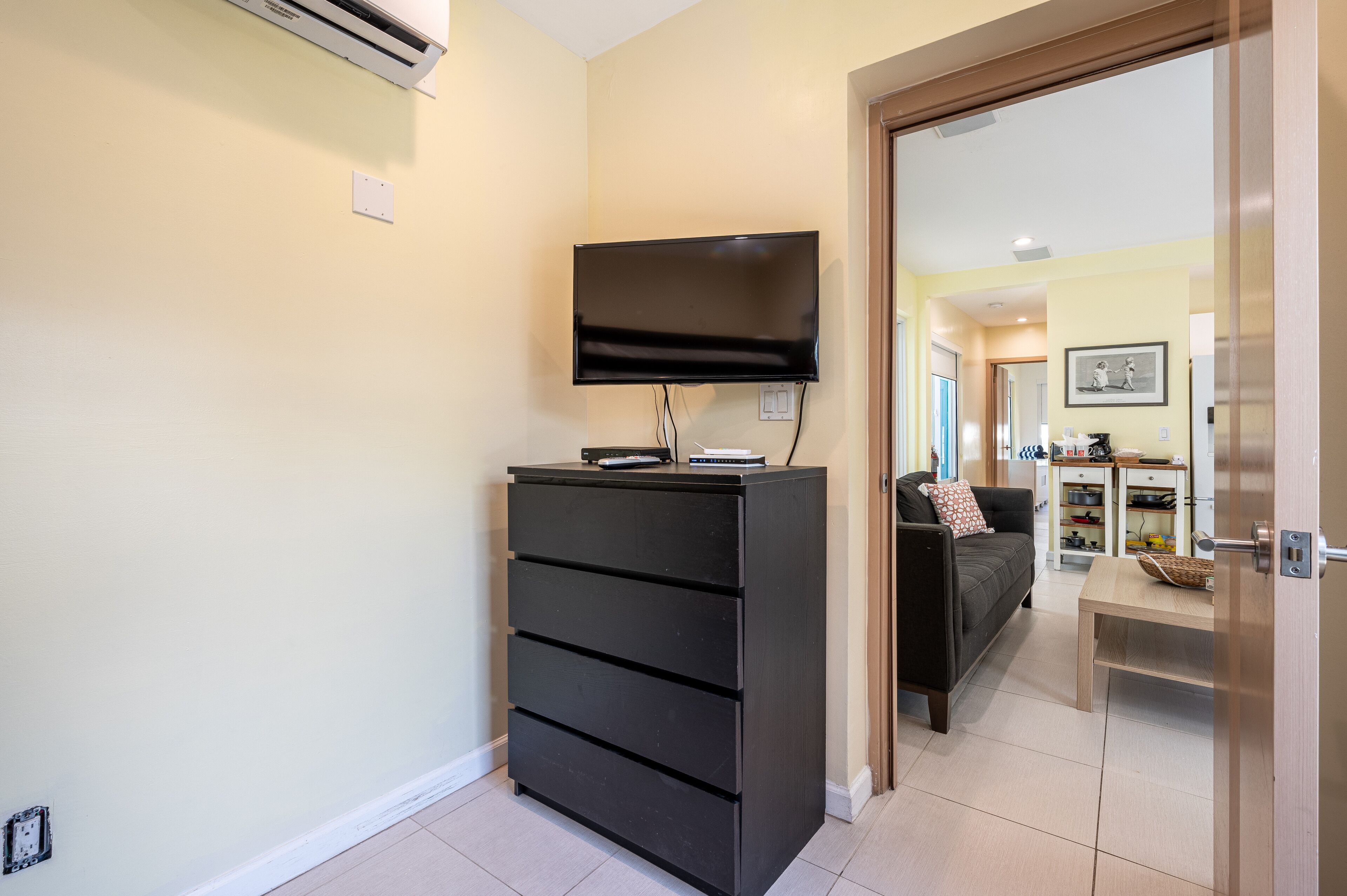 A compact living space with a flat-screen TV mounted on the wall, a black storage unit, a sofa with a patterned cushion, and a wooden table.