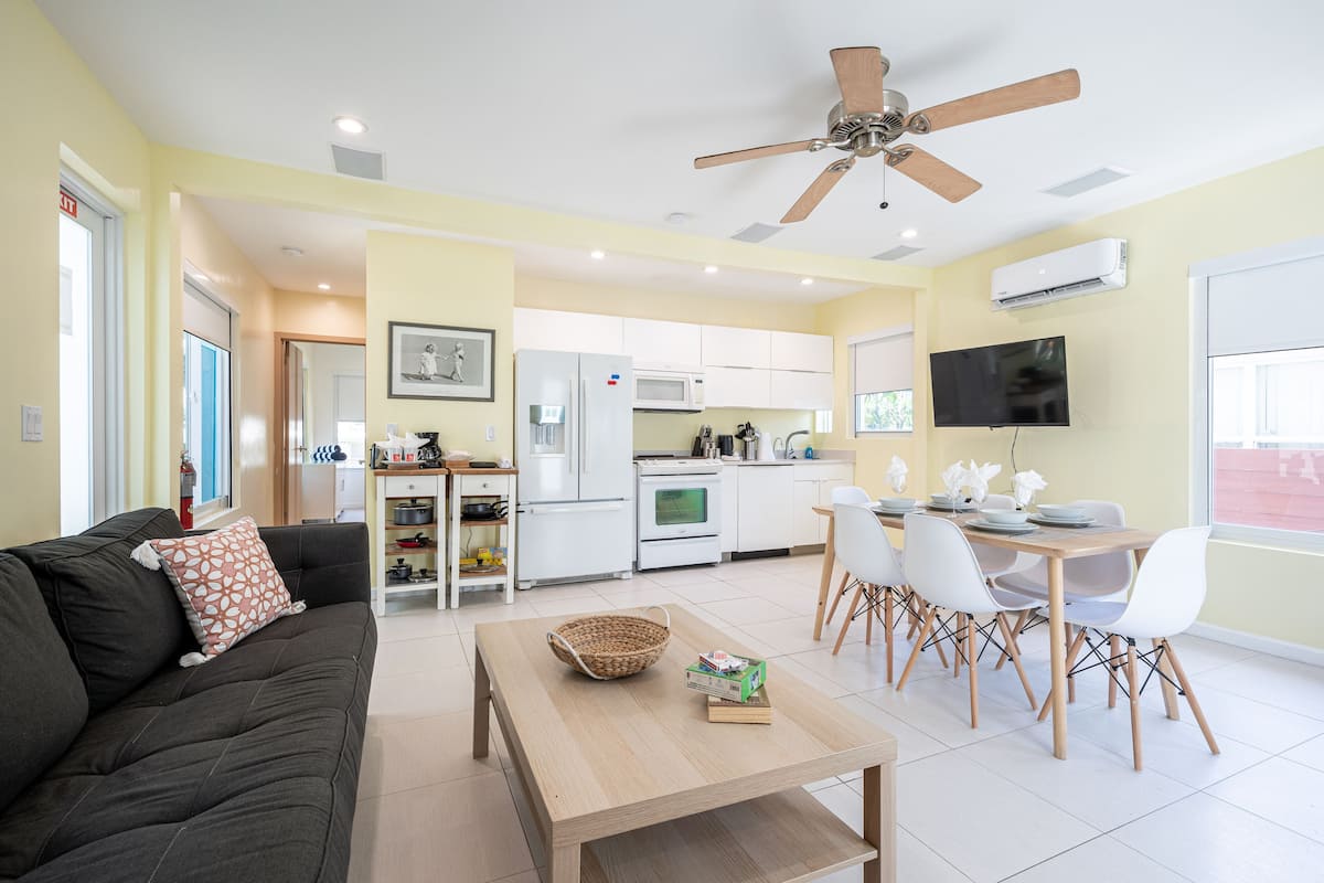 A modern living room with a sofa, a dining table with chairs, a kitchen area with appliances, and a ceiling fan.