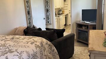 A bedroom with a large bed, a TV, and a kitchenette in the background.