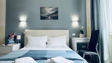 A hotel room with a bed, two pillows, a bedside table with a lamp, a chair, a small table with a plant, and a painting on the wall.