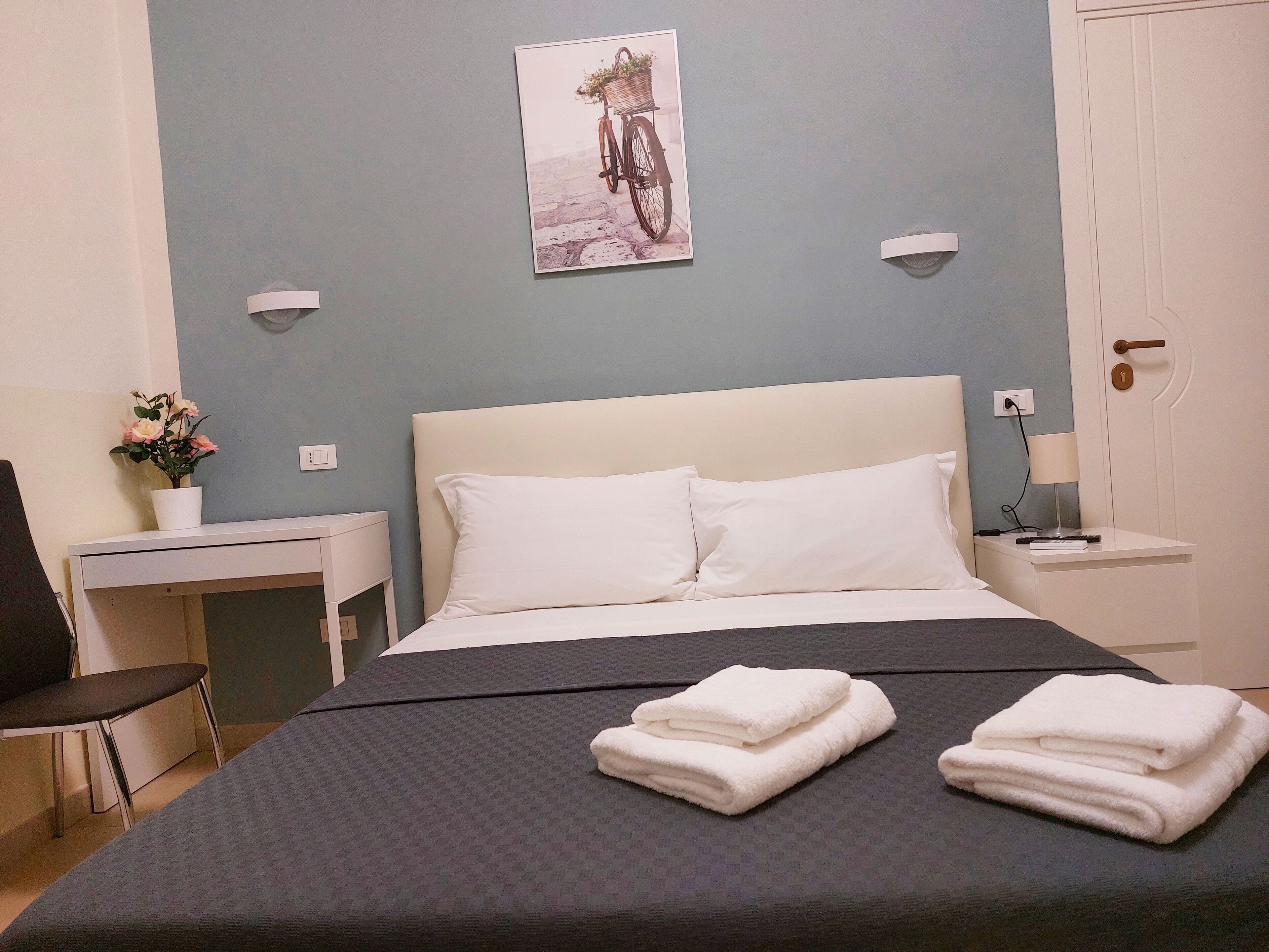 Un letto rifatto con due asciugamani piegati, una piccola scrivania con una sedia, una lampada a parete e un quadro con una bicicletta.