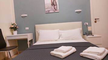 Un letto rifatto con due asciugamani piegati, una piccola scrivania con una sedia, una lampada a parete e un quadro con una bicicletta.