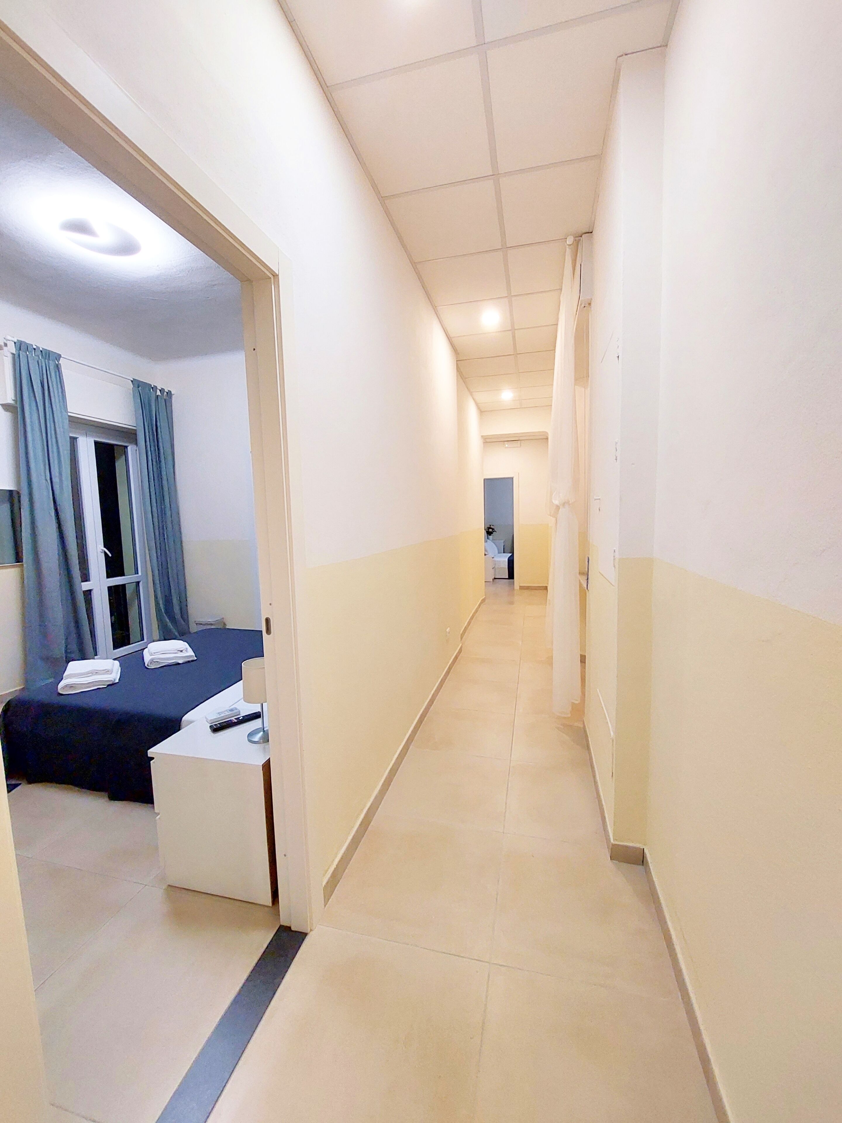 Un couloir d’hôtel avec un lit, un bureau et une salle de bain visibles au fond.