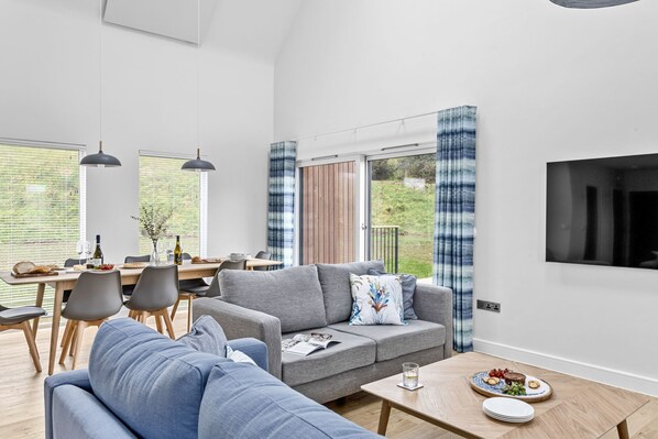 A modern living room with a sofa, a dining table, and a TV mounted on the wall. - 8 The Vista, Willingcott Nr Woolacombe - Hot tub/ On-site pool/ EV Charger/ Ecohome (Woolacombe)