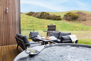A hot tub with a table and chairs, a grill, and a view of a grassy hill. - 8 The Vista, Willingcott Nr Woolacombe - Hot tub/ On-site pool/ EV Charger/ Ecohome (Woolacombe)