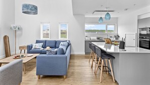 Une cuisine moderne avec un îlot central, un coin repas doté d’un canapé bleu et une vue sur l’extérieur grâce à de grandes fenêtres.