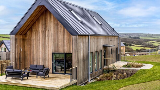 Une maison moderne en bois, dotée d’un toit à pente, de grandes fenêtres et d’une terrasse aménagée avec du mobilier d’extérieur.