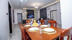 Un coin repas avec une table en bois dressée pour quatre personnes, un téléviseur fixé au mur et un canapé à l’arrière-plan.