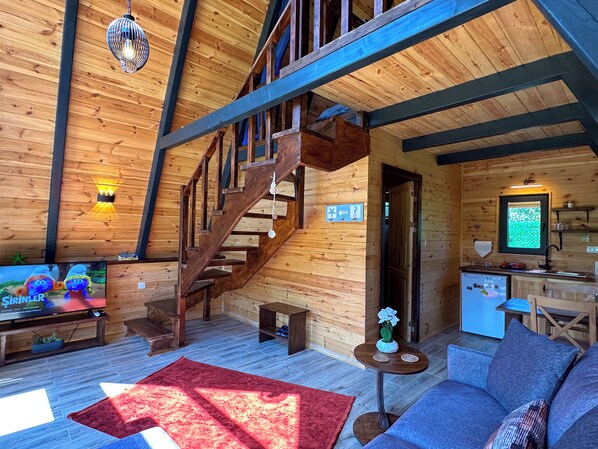 A wooden cabin interior with a staircase, a TV, a sofa, and a kitchen area.