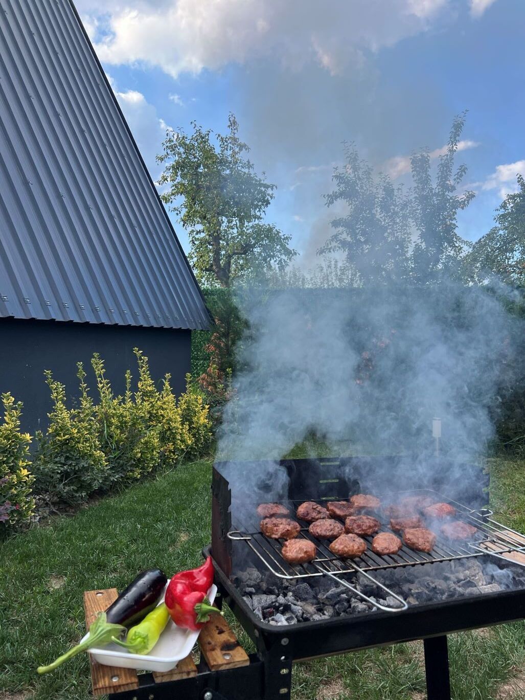 Ein Grill mit Fleisch, umgeben von viel Grün und einem Gebäude.