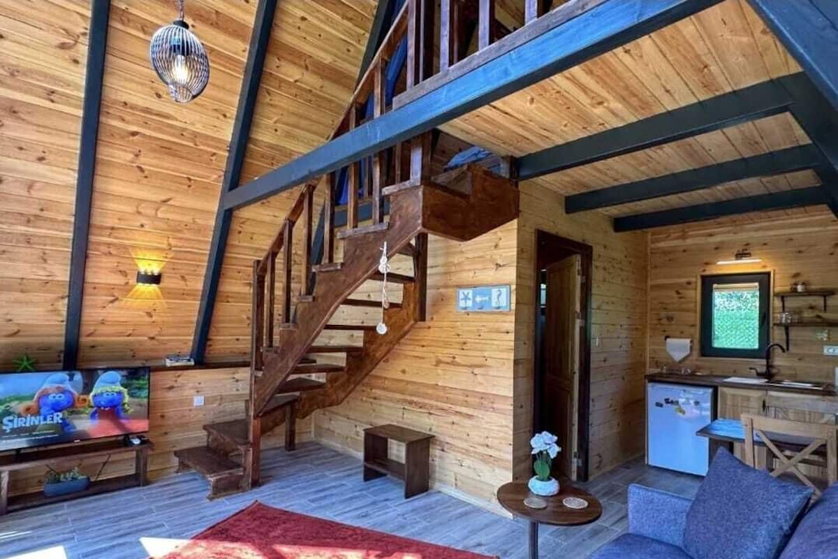 A wooden cabin interior with a staircase, a sofa, a TV, and a kitchenette.