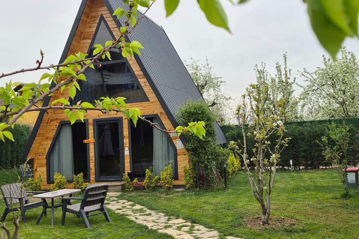 A triangular wooden cabin with a glass door, a small patio with a table and chairs, and a garden with a pathway leading to the cabin.