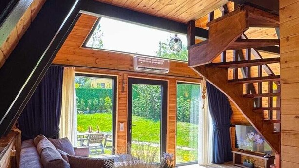 Acogedor cuarto en el ático con techo de madera, un sofá, una mesa con un florero y vistas a un jardín a través de amplias ventanas.